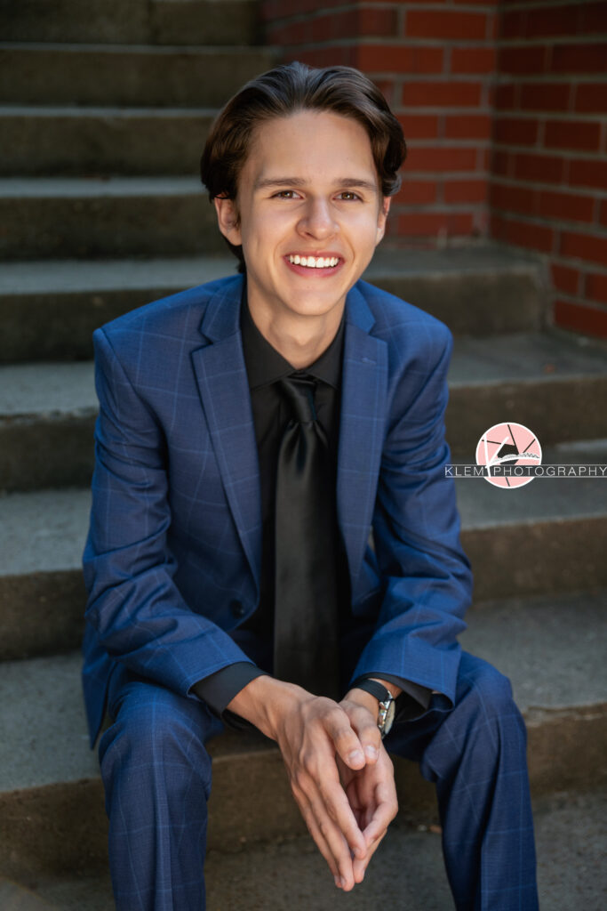 Henderson KY Senior pics with Klem Photography. Senior boy with short brown hair in blue plaid suit and pants wearing black undershirt and black tie smiles at the camera. He sits on concrete steps with red brick wall in the background. His hands sit together in front of him resting on his legs.