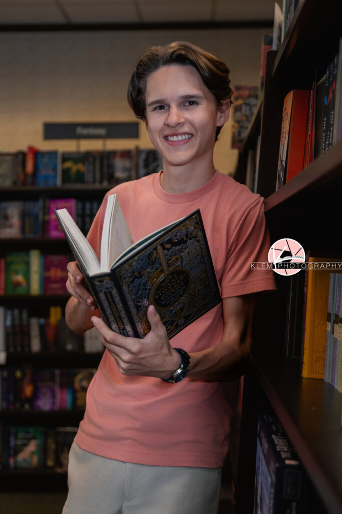 Henderson KY Senior pics with Klem Photography. Senior boy with short brown hair wearing a coral shirt and khaki pants smiles at the camera. He leans against a bookshelf and is holding a book.