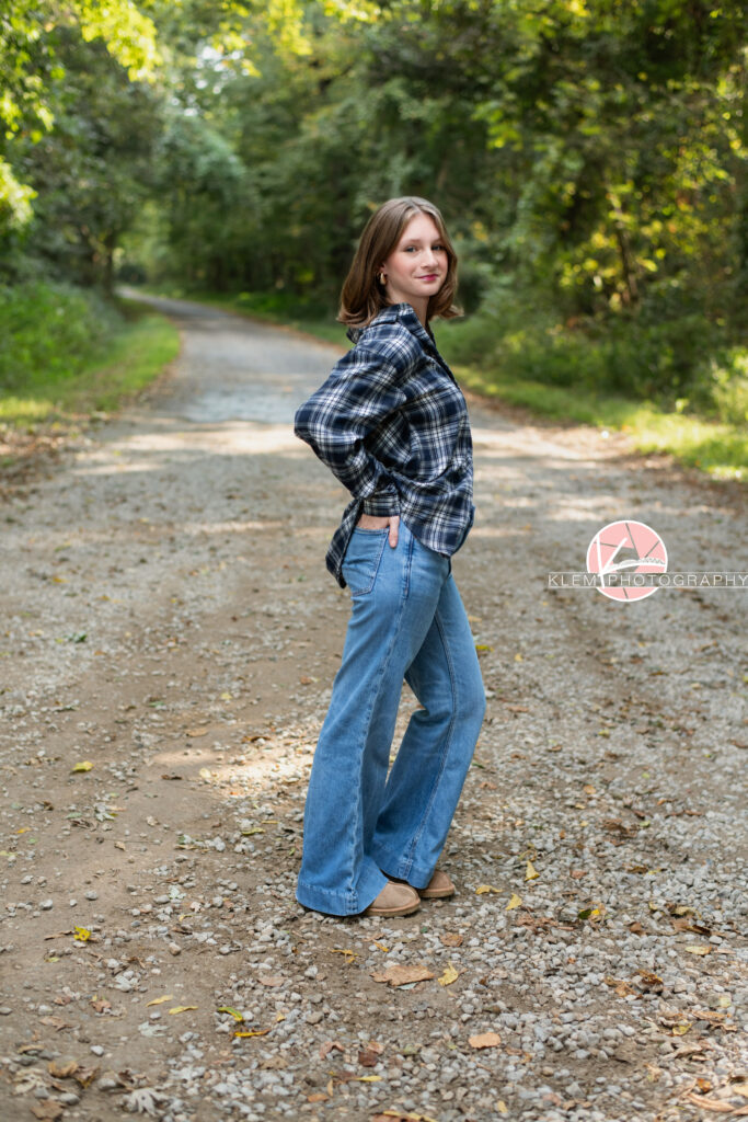 Fall Senior Pictures, Henderson KY, Klem Photography. Senior girl with medium brown hair wearing black and white flannel, jeans and brown boots smiles at the camera. she stands facing sideways with her hand resting in her pocket and one leg bent. She stands on a gravel road with trees in the background.