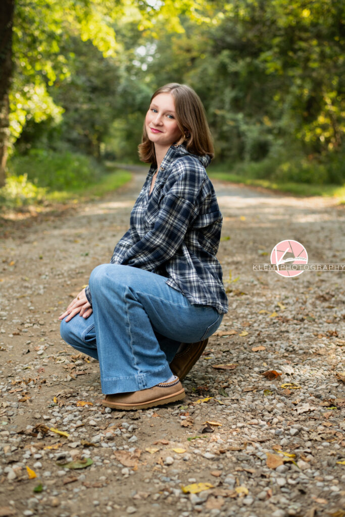 Fall Senior Pictures, Henderson KY, Klem Photography. Senior girl with medium brown hair wearing black and white flannel, jeans and brown boots smiles at the camera. She crouches with her arms resting on her legs on a gravel road with trees in the background.
