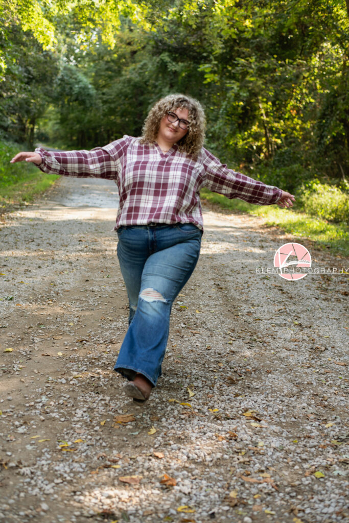 Fall Senior Pictures, Henderson KY, Klem Photography. Senior girl with long brown curly hair and glasses, wearing a red and white flannel, jeans, and brown boots smiles at the camera. She walks forward with her arms out and one leg up. She stands on a gravel road with trees in the background.