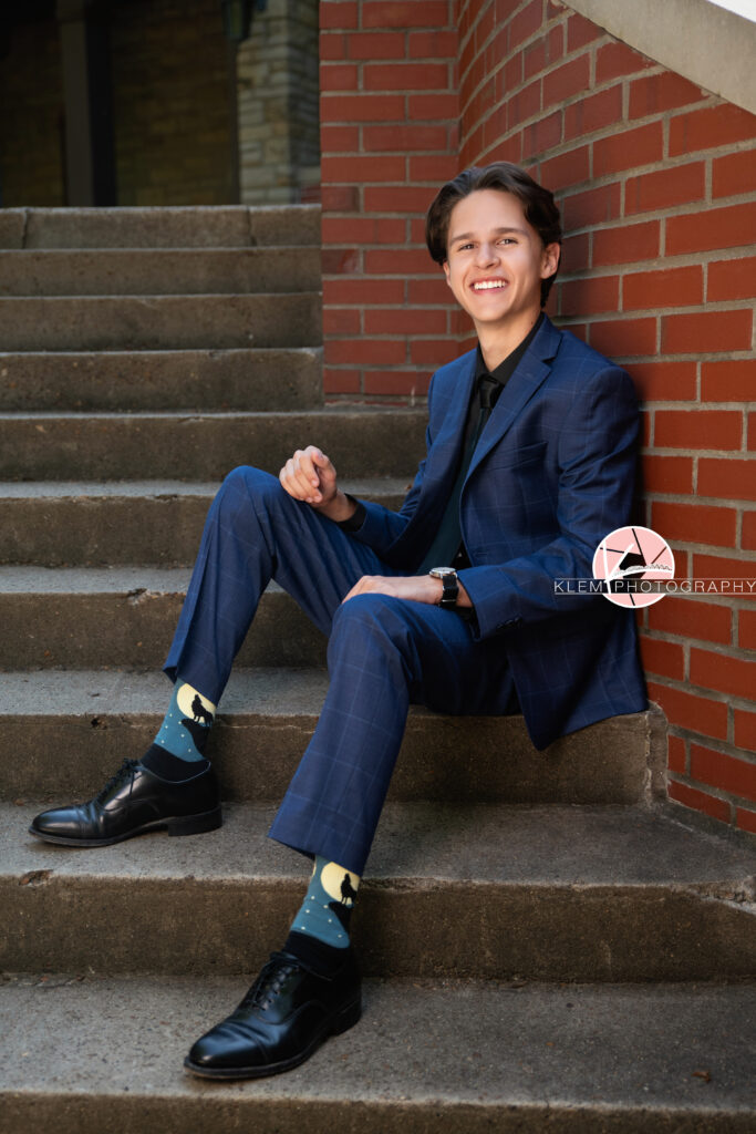Henderson KY Senior pics with Klem Photography. Senior boy with short brown hair in blue plaid suit and pants wearing black undershirt and black tie along with blue wolf socks and black dress shoes smiles at the camera. He sits on concrete steps leaning against a red brick wall and each arm rests on one of his legs.
