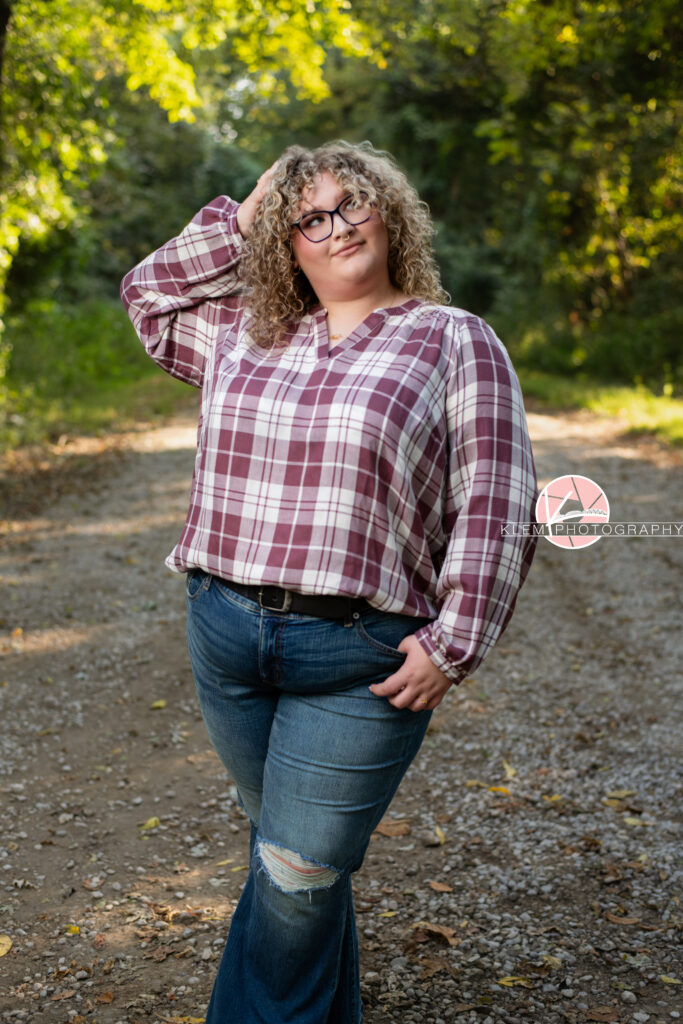 Fall Senior Pictures, Henderson KY, Klem Photography. Senior girl with long brown curly hair and glasses, wearing a red and white flannel, jeans, and brown boots smiles at the camera. She looks up and off to the right of the camera with one hand in her hair and the other in her front pocket. She stands with her legs crossed on a gravel road with trees in the background.
