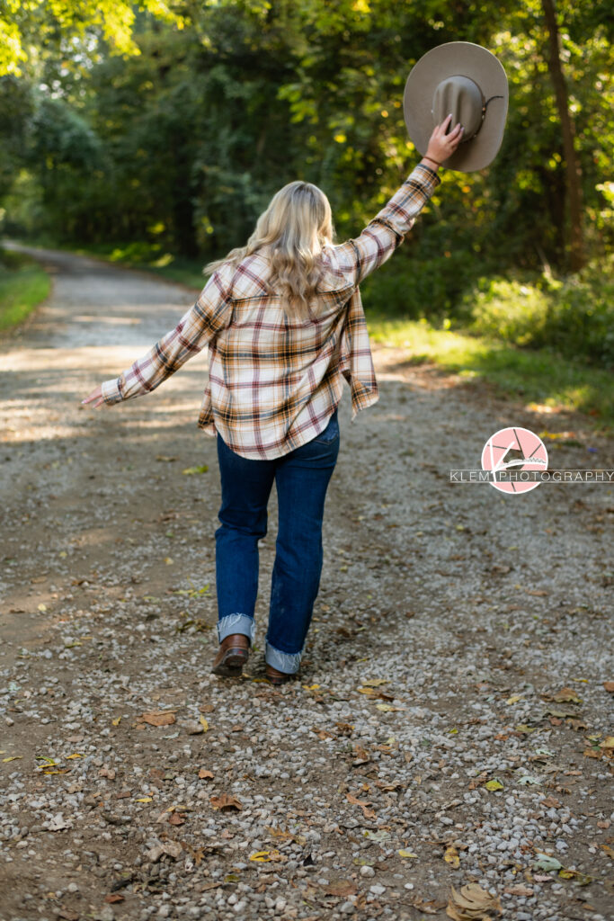 Fall Senior Pictures, Henderson KY, Klem Photography. Senior girl with long blond hair wearing red, tan, and white flannel, jeans, and brown boots faces away from the camera. She holds a cowboy hat above her head with one arm out as she walks away from the camera.