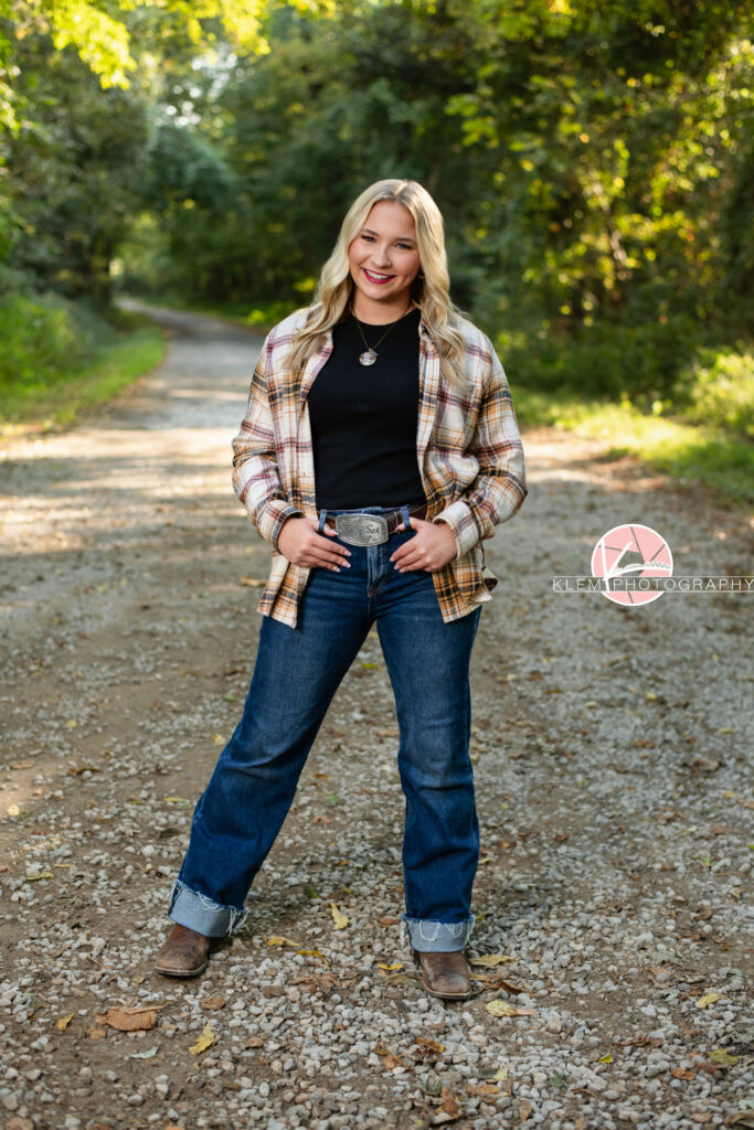 Fall Senior Pictures, Henderson KY, Klem Photography. Senior girl with long blond hair wearing red, tan, and white flannel over black shirt, jeans, a belt with a silver belt buckle, and brown boots smiles at the camera. She stands with her legs apart and one hip popped with her hands in her belt loops. She stands on a gravel road with trees in the background.