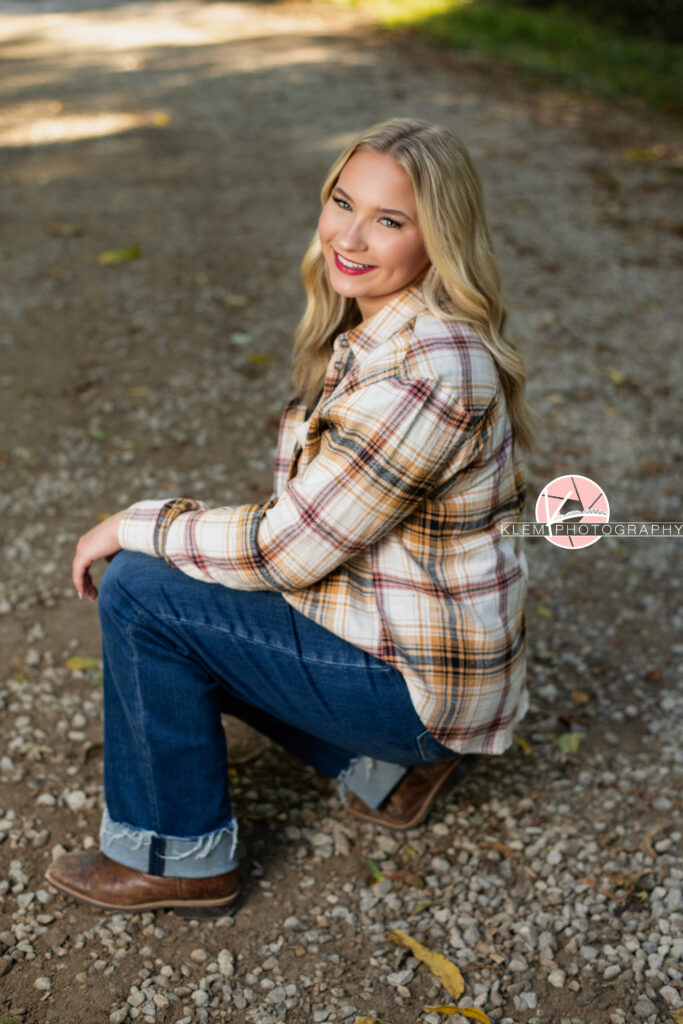 Fall Senior Pictures, Henderson KY, Klem Photography. Senior girl with long blond hair wearing red, tan, and white flannel, jeans, and brown boots smiles at the camera. Her arm rests on her leg. She crouches on a gravel road smiling at the camera.