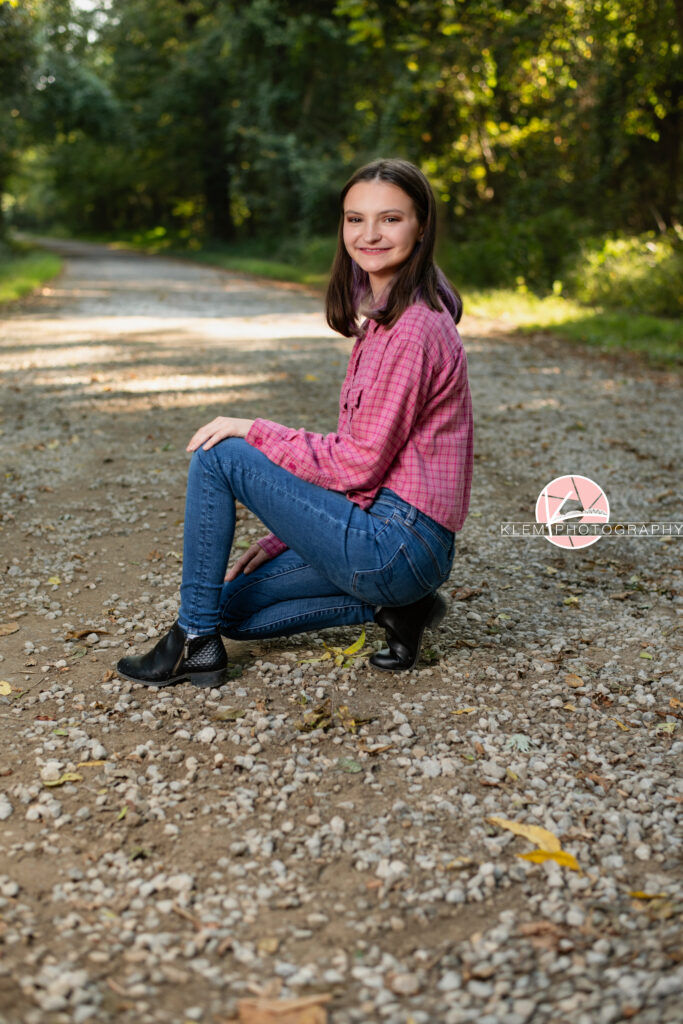 Fall Senior Pictures, Henderson KY, Klem Photography. Senior girl with medium brown hair wearing red flannel, jeans and black boots smiles at the camera. She crouches facing sideways with her arms resting on her legs. She is on a gravel road with trees in the background.