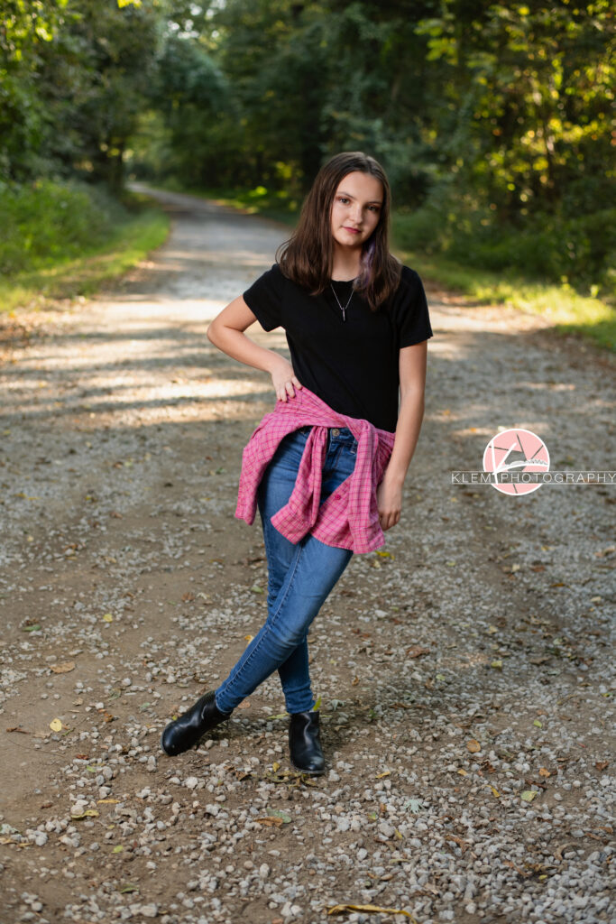 Fall Senior Pictures, Henderson KY, Klem Photography. Senior girl with medium brown hair wearing red flannel around her waist, black shirt, jeans and black boots looks straight faced at the camera. She has her legs crossed and rests one arm on her hip. She stands on a gravel road with trees in the background.