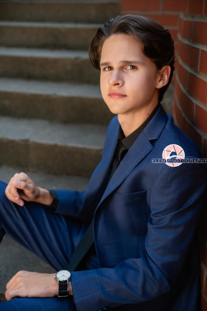 Henderson KY Senior pics with Klem Photography. Senior boy with short brown hair in blue plaid suit and pants wearing black undershirt and black tie looks straight-faced at the camera. He sits on concrete steps with a red brick wall in the background and each arm rests on one of his legs.