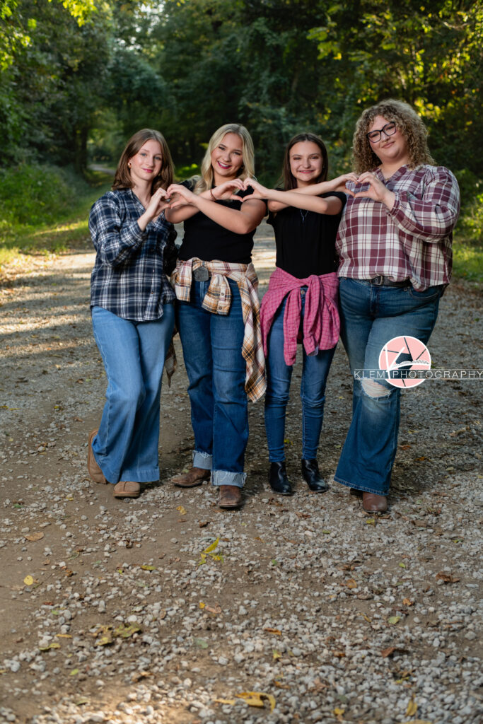 Fall Senior Pictures, Henderson KY, Klem Photography. From left to right: Senior girl with medium brown hair with black and white flannel, jeans, and brown boots smiles at the camera and makes a heart with half of her hand with the girl next to her. Senior girl with long blond hair wearing white, red, and brown flannel  around her waist over black shirt with jeans and brown boots smiles at the camera. She completes the previous heart with her hand and makes another half of a heart with the girl next to her. Senior girl with medium brown hair wearing red flannel around her waist over a black shirt with jeans and black boots smiles at the camera. She completes the previous heart with her hands and makes another half of a heart with the girl next to her. Senior girl with long brown curly hear wearing glasses, a red and white flannel, jeans, and brown boots smiles at the camera. She completes the previous heart with her hand. They all stand on a gravel road with trees in the background.