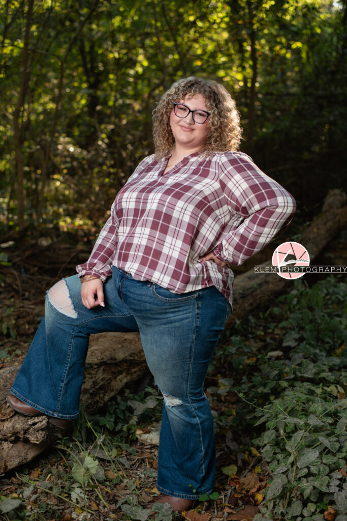 Fall Senior Pictures, Henderson KY, Klem Photography. Senior girl with long brown curly hair and glasses, wearing a red and white flannel, jeans, and brown boots smiles at the camera. She stands with one leg up on a log with one hand on her hip and the other on her leg. She stands in a forest with greenery in the background.