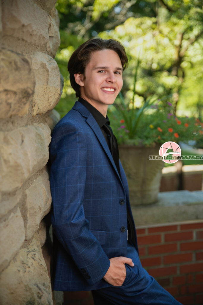 Henderson KY Senior pics with Klem Photography. Senior boy with short brown hair in blue plaid suit and pants wearing black undershirt and black tie smiles at the camera. He leans against a rock wall and has one hand in his pocket. Greenery is visible in the background.