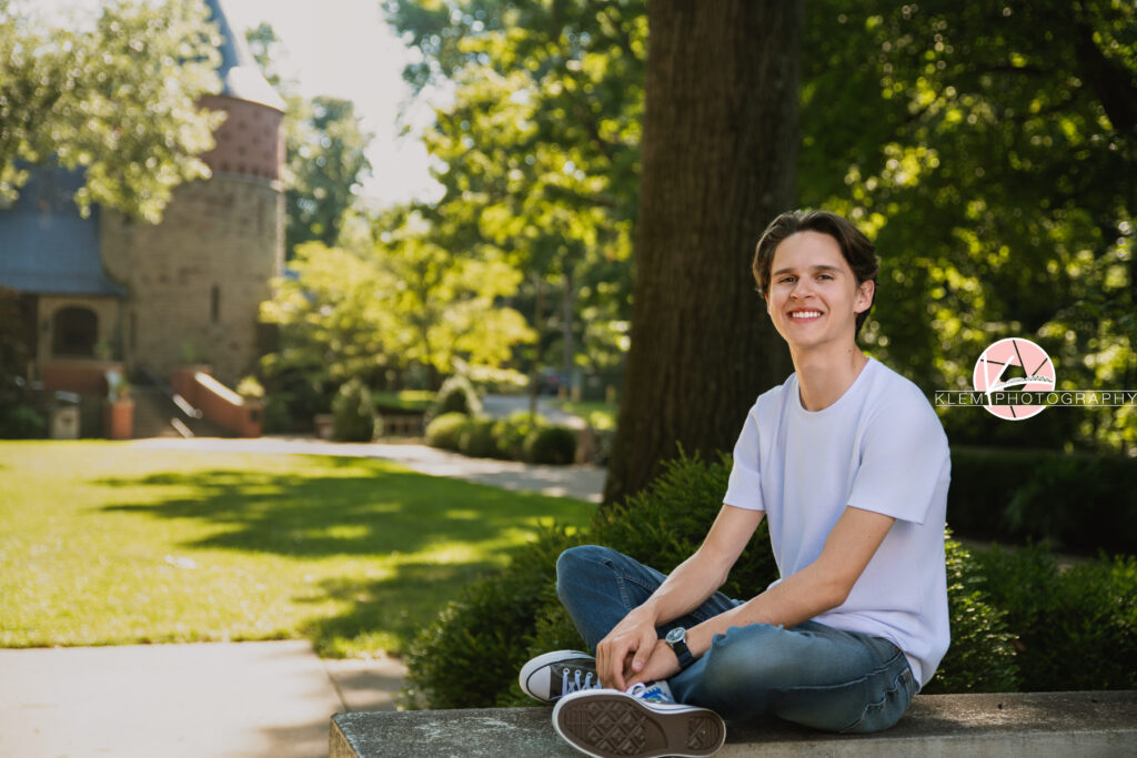 Henderson KY Senior pics with Klem Photography. Senior boy with short brown hair wearing a white shirt, jeans, and grey converse smiles at the camera. He sits with his legs crossed on a concrete railing and his arms rest on his legs. A castle and greenery are visible in the background.