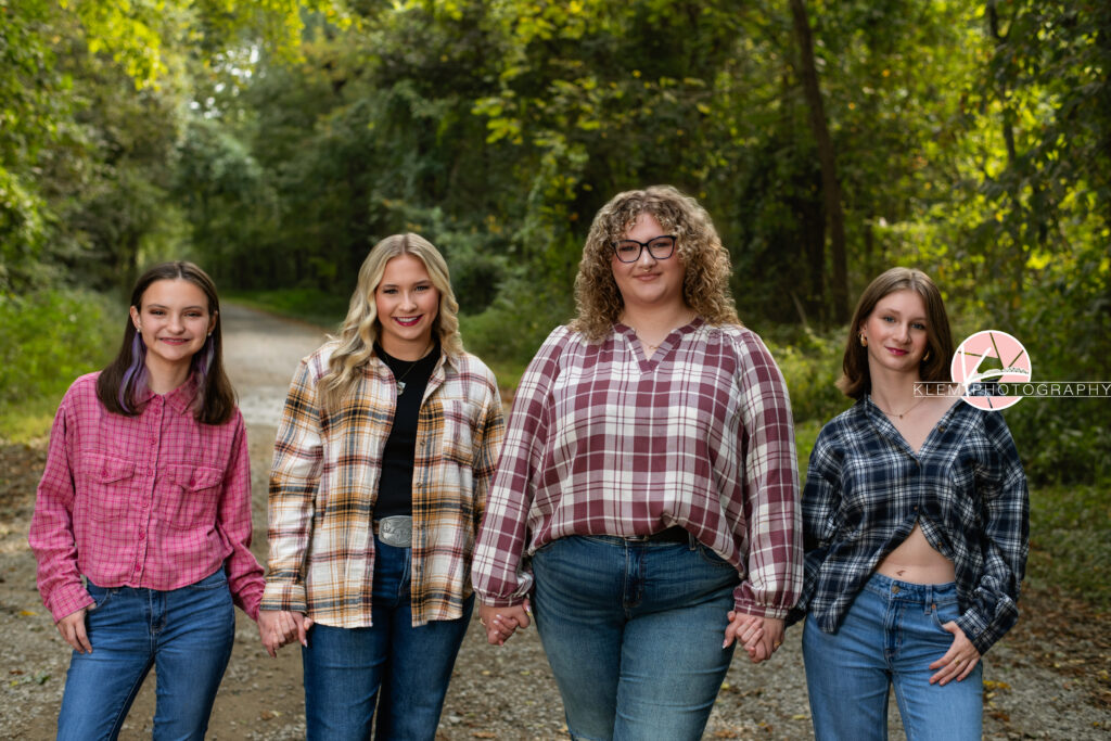 Fall Senior Pictures, Henderson KY, Klem Photography. From left to right: Senior girl with medium brown hair with purple underneath wearing red flannel and jeans smiles at the camera. She stands with one hand in her front pocket and the other holding hands with the girl next to her. Senior girl with long blond hair wearing red, white, and tan flannel over a black shirt, jeans, and a belt with a silver belt buckle smiles at the camera. She holds hands with the girls on either side of her. Senior girl with long curly brown hair, glasses, a red and white flannel. and jeans smiles at the camera. She holds hands with the girls on either side of her. Senior girl with medium brown hair wearing black and white flannel and jeans smiles at the camera. She stands with one hand in her pocket and the other hand holding the girl next to hers hand. They all stand on a gravel road with trees in the background.