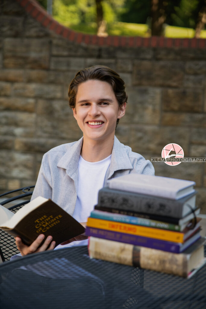 Henderson KY Senior pics with Klem Photography. Senior boy with short brown hair in a grey button up over a white shirt smiles at the camera. He sits in a wire chair at a table holding an open book while more books rest on the table. A rock wall and trees are visible in the background.
