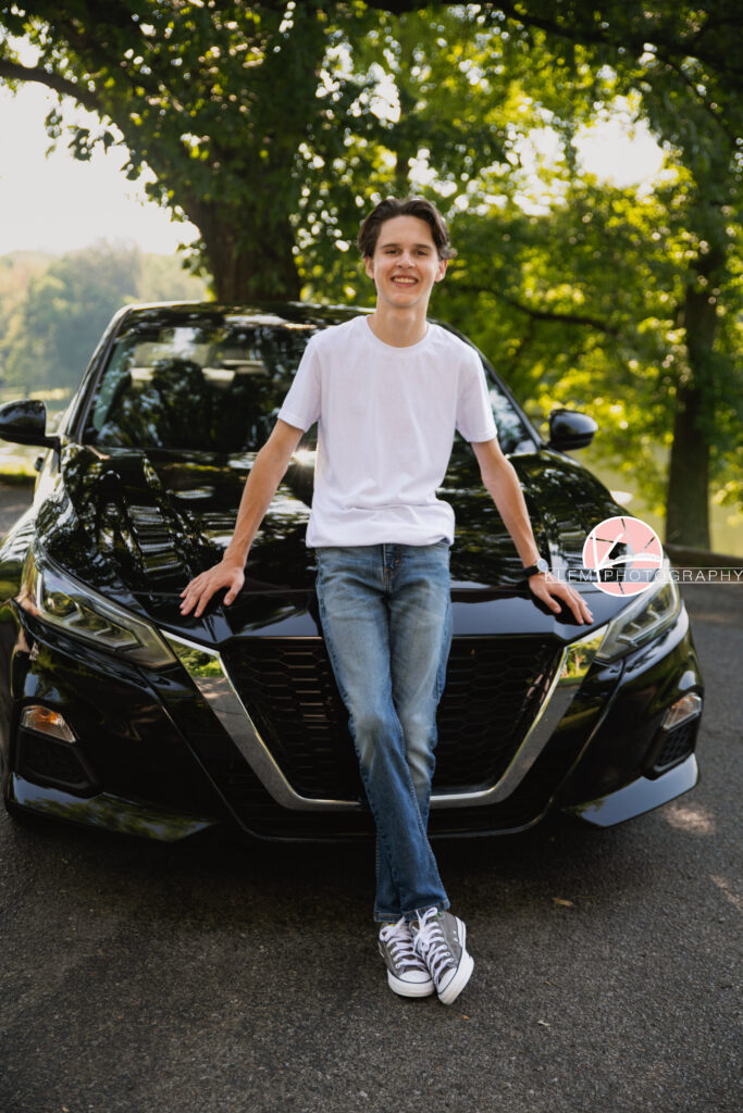 Henderson KY Senior pics with Klem Photography. Senior boy with short brown hair wearing a white shirt, jeans, and grey converse smiles at the camera. He leans against a black car with his legs crossed and trees are visible in the background.