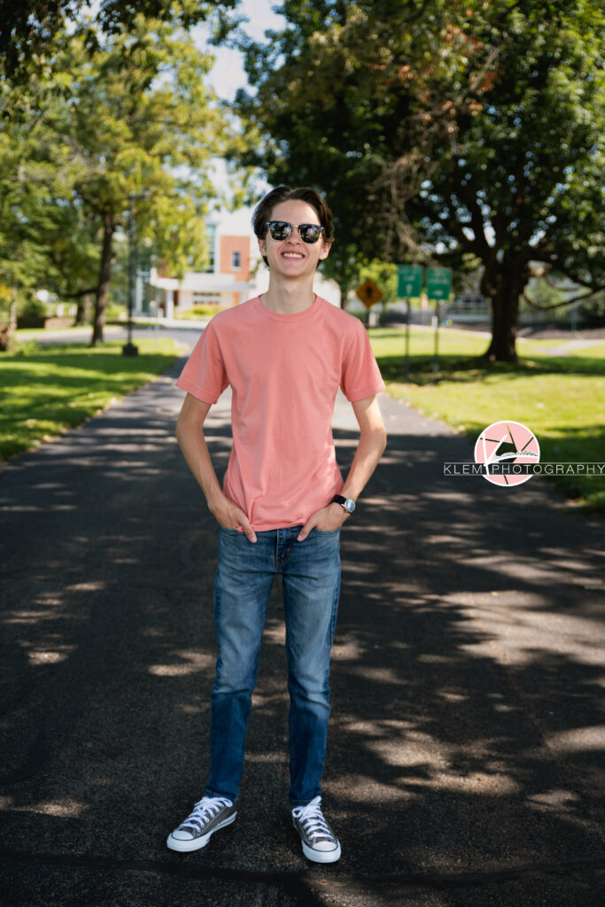 Henderson KY Senior pics with Klem Photography. Senior boy with short brown hair wearing a coral shirt, jeans, grey converse, and sunglasses smiles at the camera. he stands with his hands in his pockets on a road with trees visible in the background.