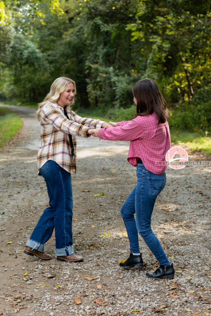 Fall Senior Pictures, Henderson KY, Klem Photography. Two senior girls, one with long blond hair, wearing white, red, and tan flannel, jeans, and brown boots smiling at the camera holds hands with senior girl with medium brown hair, wearing red flannel, jeans, and black boots. They are spinning together on a gravel road with trees in the background.