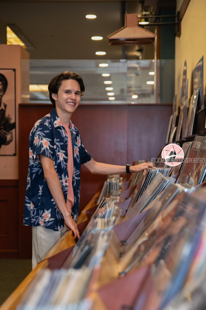 Henderson KY Senior pics with Klem Photography. Senior boy with short brown hair wearing a blue floral button up over a coral shirt and khaki pants smiles at the camera. He stands in front of a display of books and his hands rest on the books. 