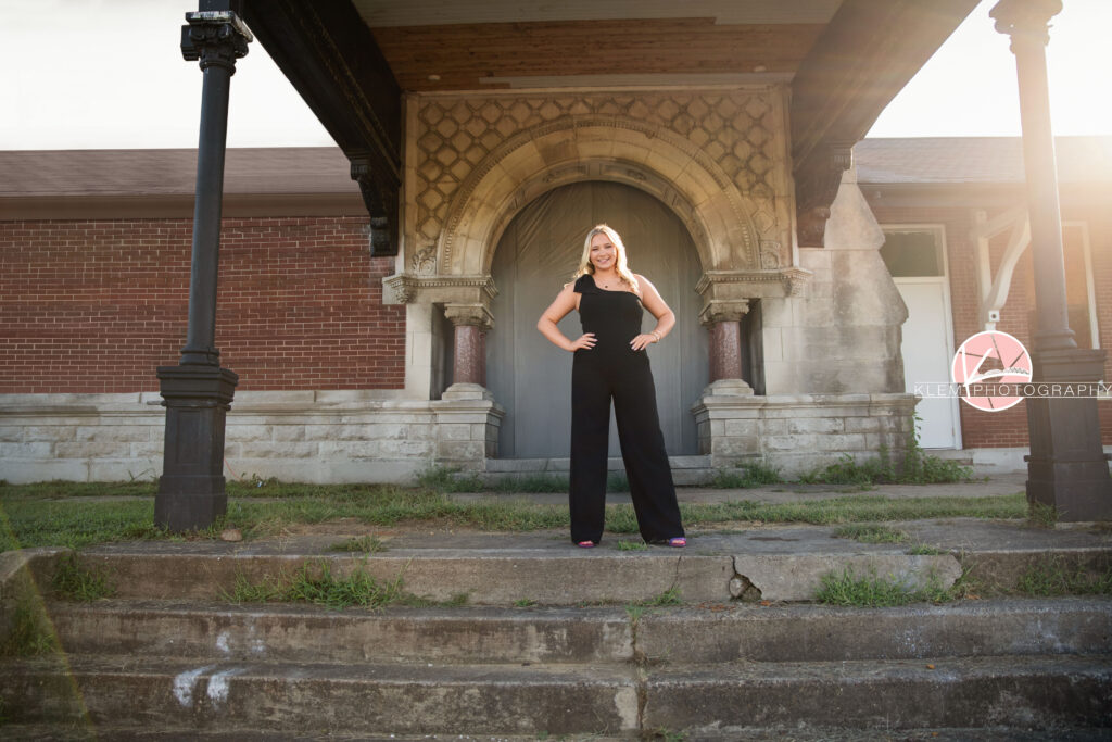 Senior Pictures Henderson KY, Avery, Webster County High School, Klem Photography. Full body shot of senior girl with long blond hair wearing a black jumpsuit with one shoulder and pink heels smiles at the camera with her hands on her hips. She is standing at the top of concrete steps with grass poking through with columns and an archway behind her.