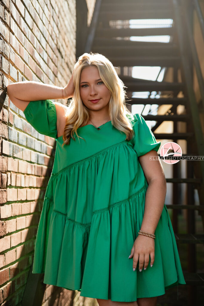 Senior Pictures Henderson KY, Avery, Webster County High School, Klem Photography. Half body shot of senior girl with long blond hair wearing a green dress and black sandals looks at the camera. She stands with one hand in her hair and one hand by her side leaning on a brick wall with a metal staircase in the background.