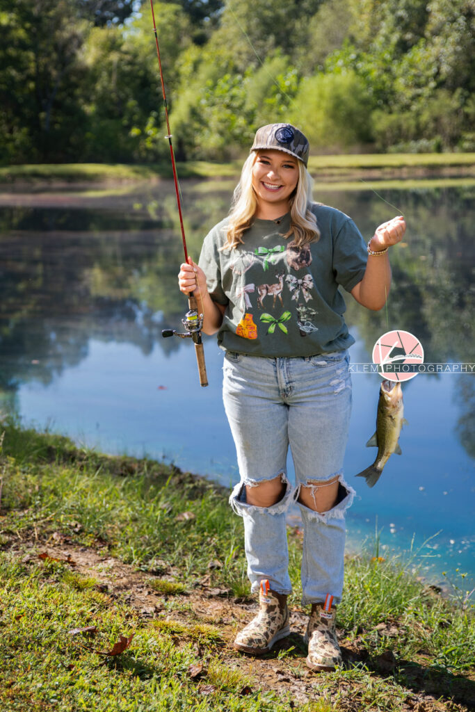 Senior Pictures Henderson KY, Avery, Webster County High School, Klem Photography. Full body shot of senior girl with long blond hair wearing a shirt with bows and deer, ripped light wash blue jeans, a camo hat, and boots smiles at the camera. She stands holding a fishing pole with a fish on it with a pond and trees in the background.