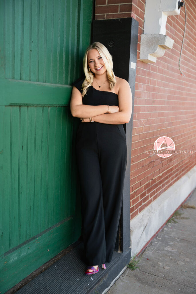 Senior Pictures Henderson KY, Avery, Webster County High School, Klem Photography. Full body shot of senior girl with long blond hair wearing a black jumpsuit with one shoulder and pink heels smiles at the camera. She stands with her arms crossed and one foot in front of the other leaning against a doorway with a green door, brick wall, and concrete sidewalk visible around her.
