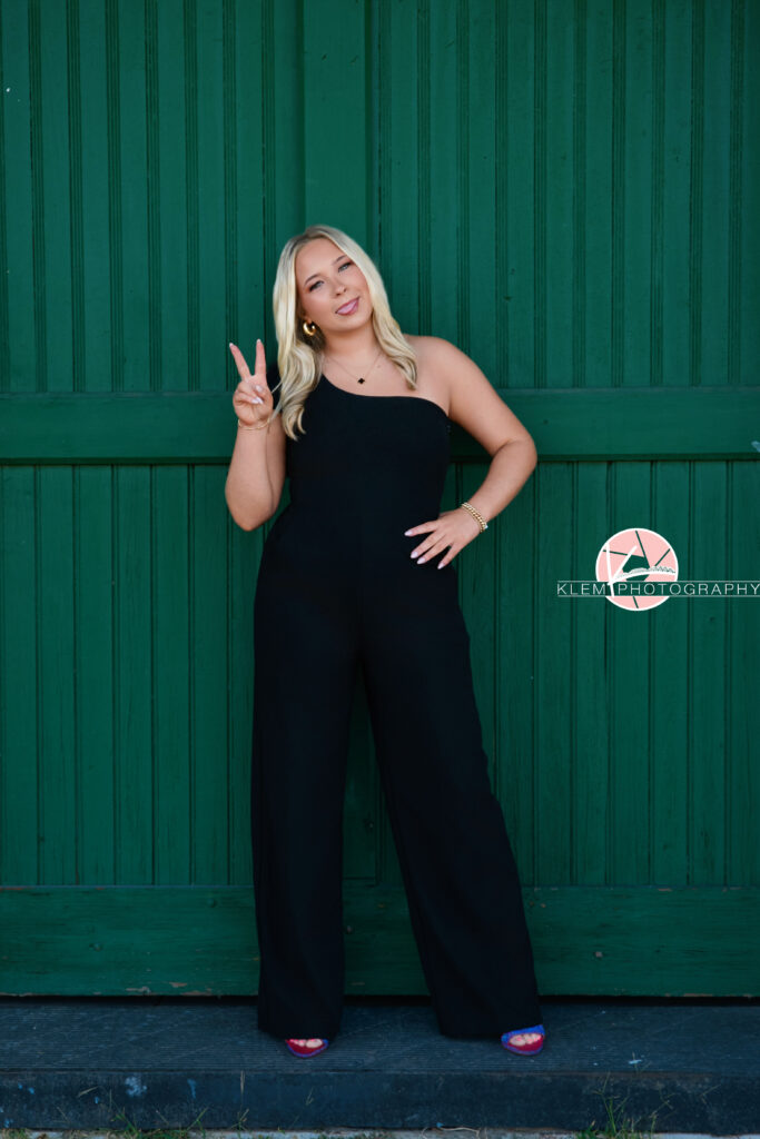 Senior Pictures Henderson KY, Avery, Webster County High School, Klem Photography. Full body shot of senior girl with long blond hair wearing a black jumpsuit with one shoulder and pink heels makes a silly face at the camera. She stands with her legs apart holding up a peace sign with one hand, sticking her tongue out, and with one hand on her hip. There is a green door visible behind her.