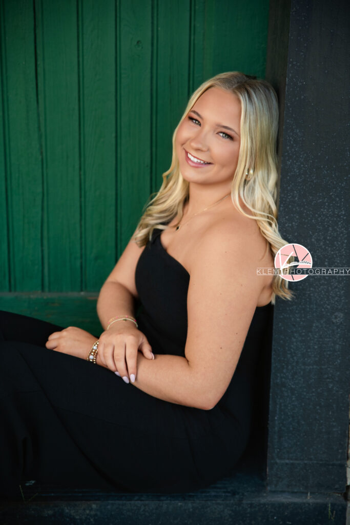 Senior Pictures Henderson KY, Avery, Webster County High School, Klem Photography. Half body shot of senior girl with long blond hair wearing a black jumpsuit with one shoulder smiles at the camera. She sits with her arms on her lap facing sideways. She sits leaning against a wall with a green door in the background.