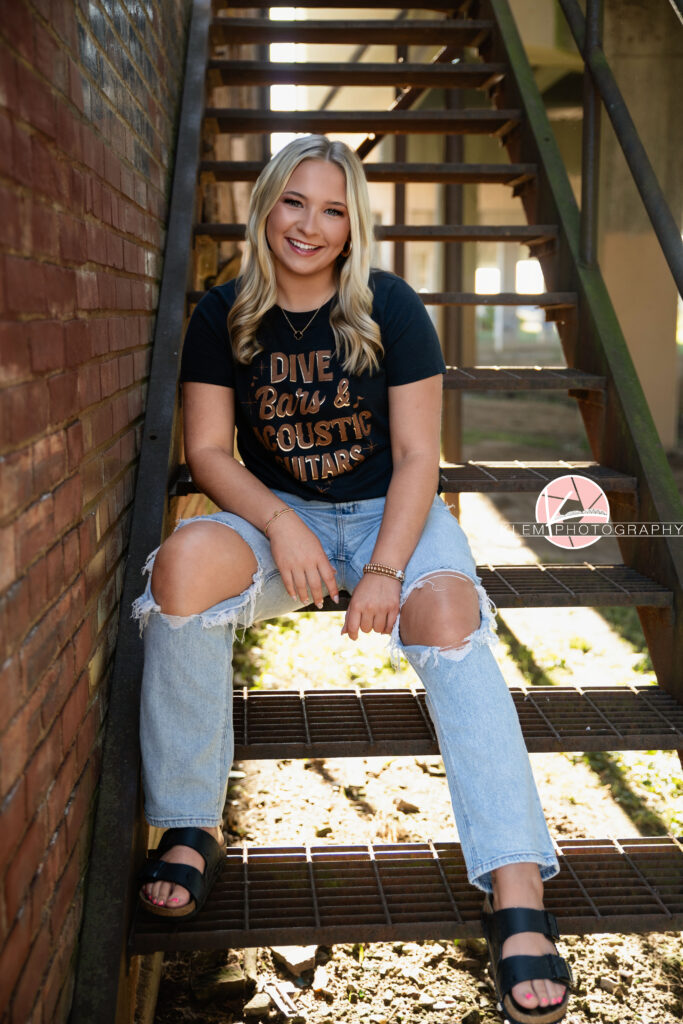 Senior Pictures Henderson KY, Avery, Webster County High School, Klem Photography. Full body shot of senior girl with long blond hair wearing a dive bar shirt, ripped light wash blue jeans, and sandals smiles at the camera. She sits with her hands resting on her legs on a metal staircase by a brick wall and concrete pillars in the background.