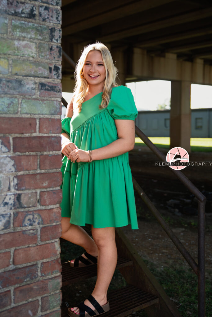 Senior Pictures Henderson KY, Avery, Webster County High School, Klem Photography. Full body shot of senior girl with long blond hair wearing a green dress and black sandals smiles at the camera. She stands on a metal staircase with her hands together with a brick wall to her side and concrete pillars in the background.