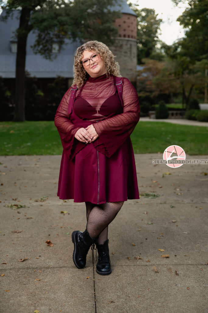 Senior Pictures, Henderson KY, Cadence, Klem Photography. Full body shot of senior girl with light brown curly hair and glasses wearing a maroon dress, black tights, and black boots smiles at the camera. She stands with her legs crossed and holding her hands together with a castle and greenery in the background.