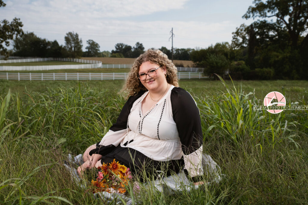 Senior Pictures, Henderson KY, Cadence, Klem Photography. half body shot of senior girl with light brown curly hair and glasses wearing a black and white dress smiles at the camera. She sits in a field with a bouquet of flowers in front of her and her legs to the side with one arm on the ground and the other on her legs with the field, a white fence, and trees in the background.