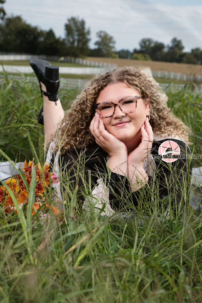 Senior Pictures, Henderson KY, Cadence, Klem Photography.  Senior girl with light brown curly hair, a nose ring, and glasses wearing a black and white dress and black boots smiles at the camera. She lays on her stomach in a field with one leg up in the background and her head resting on her hands with a bouquet next to her and the field, white fence, and trees in the background.