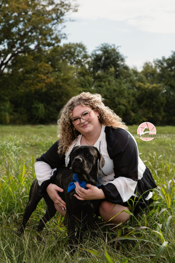 Senior Pictures, Henderson KY, Cadence, Klem Photography. Full body shot of senior girl with light brown curly hair, a nose ring, and glasses, wearing a black and white dress smiles at the camera. She crouches in a field with her arms around her black brindle cane corso dog wearing a blue bow with the field and trees in the background.