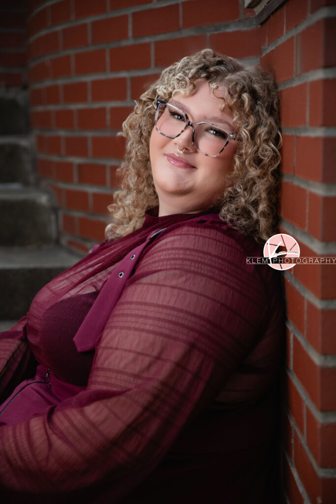Senior Pictures, Henderson KY, Cadence, Klem Photography. Half body shot of senior girl with light brown curly hair, glasses, and a nose ring wearing a maroon dress smiles at the camera. She leans against a brick wall with concrete stairs in the background.