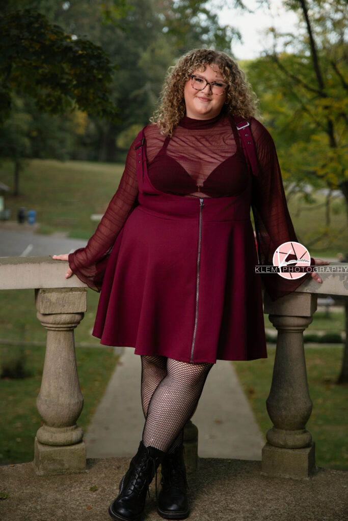 Senior Pictures, Henderson KY, Cadence, Klem Photography. Full body shot of senior girl with light brown curly hair and glasses wearing a maroon dress. black tights, and black boots smiles at the camera. She leans against a concrete landing with her arms up on it and greenery in the background.