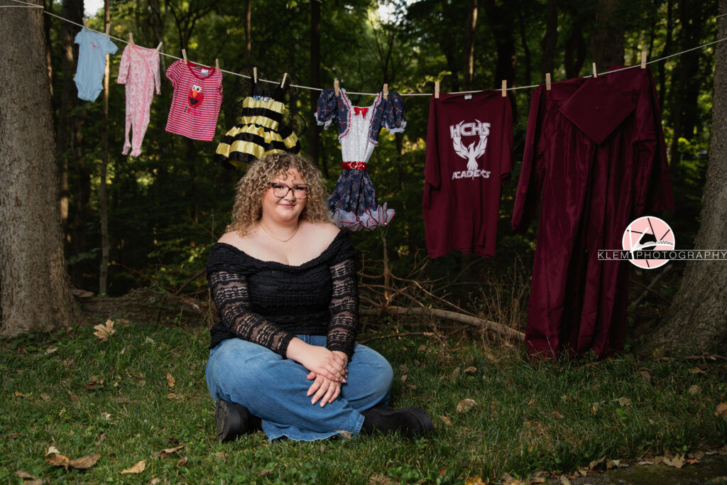 Senior Pictures, Henderson KY, Cadence, Klem Photography. Full body shot of senior girl with light brown curly hair and glasses, wearing a necklace. black ruffled long sleeve top, jeans, and black boots smiles at the camera. She sits with her legs crossed holding her arms in front of her in the grass with a clothesline with various outfits hanging between two trees in front of a forest behind her.