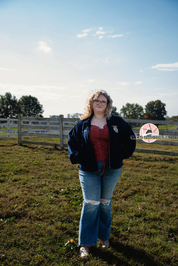 Senior Pictures, Henderson KY, Cadence, Klem Photography. Full body shot of senior girl with light brown curly hair and glasses wearing a necklace, red top, an FFA jacket, jeans and brown cowboy boots smiles at the camera. She stands with her hands in her jacket pockets walking towards the camera with a white fence and pasture behind her.
