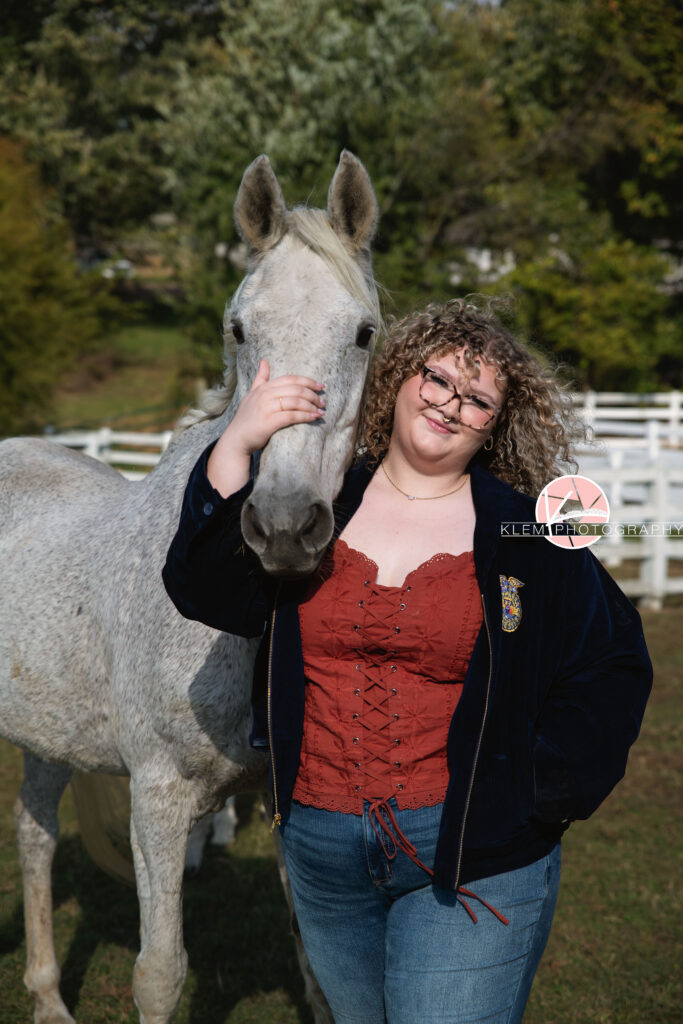 Senior Pictures, Henderson KY, Cadence, Klem Photography. Full body shot of senior girl with light brown curly hair and glasses, wearing a necklace, red top, FFA jacket, and jeans smiles at the camera with grey horse next to her. She stands with one hand on the horses face and the other in her jacket pocket with a white fence and trees in the background.