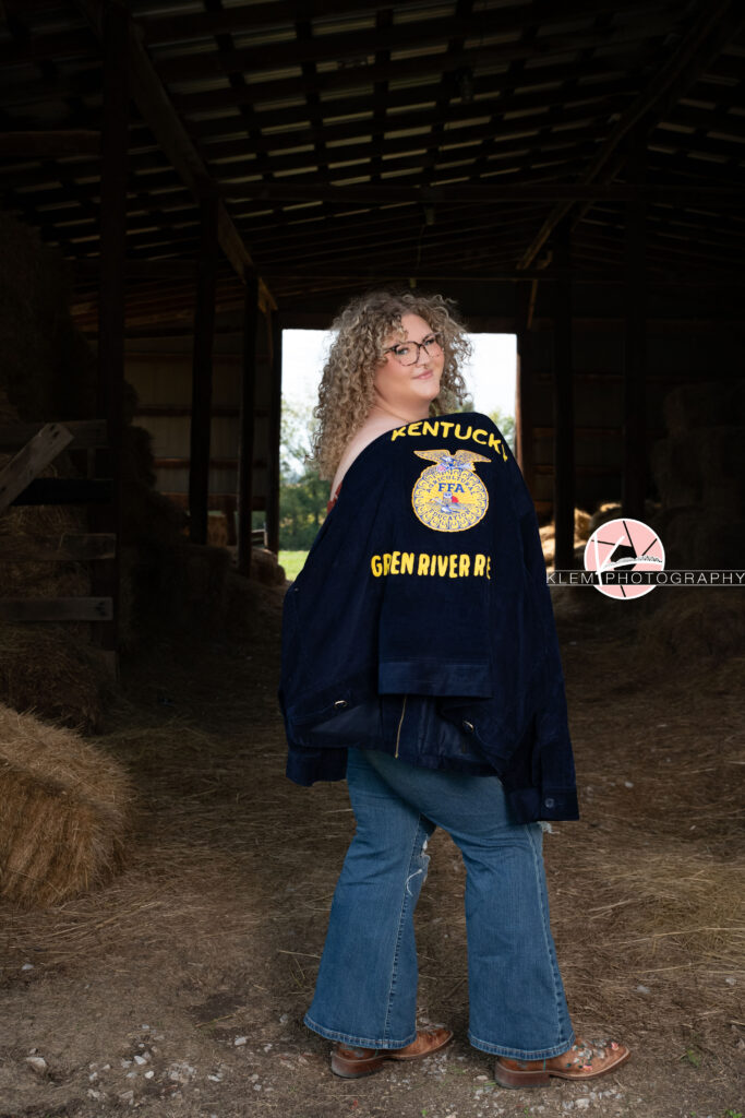 Senior Pictures, Henderson KY, Cadence, Klem Photography. Full body shot of senior girl with light brown curly hair and glasses stands holding a regional FFA jacket over her front wearing jeans and brown cowboy boots smiling at the camera. There is the inside of a barn in the background.