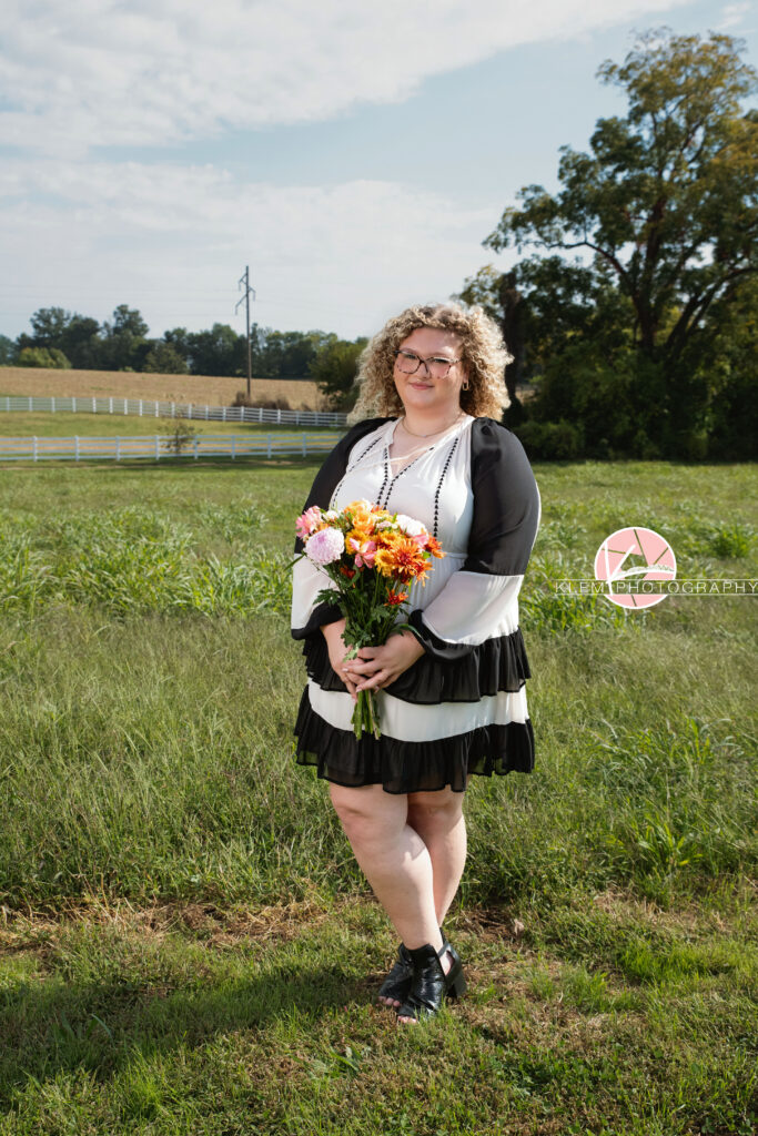 Senior Pictures, Henderson KY, Cadence, Klem Photography. Full body shot of senior girl with light brown curly hair and glasses, wearing a black and white dress and black boots smiles at the camera. She stands holding a bouquet of flowers in a field with a white fence and trees in the background.