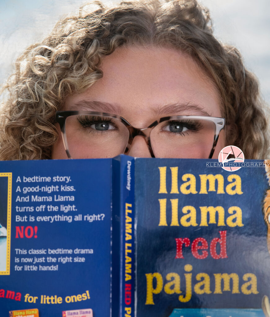 Senior Pictures, Henderson KY, Cadence, Klem Photography. Headshot of senior girl with light brown curly hair, blue eyes, and glasses looks at the camera. Half of her face is visible and the other half is covered by the cover of the llama llama red pajama book.