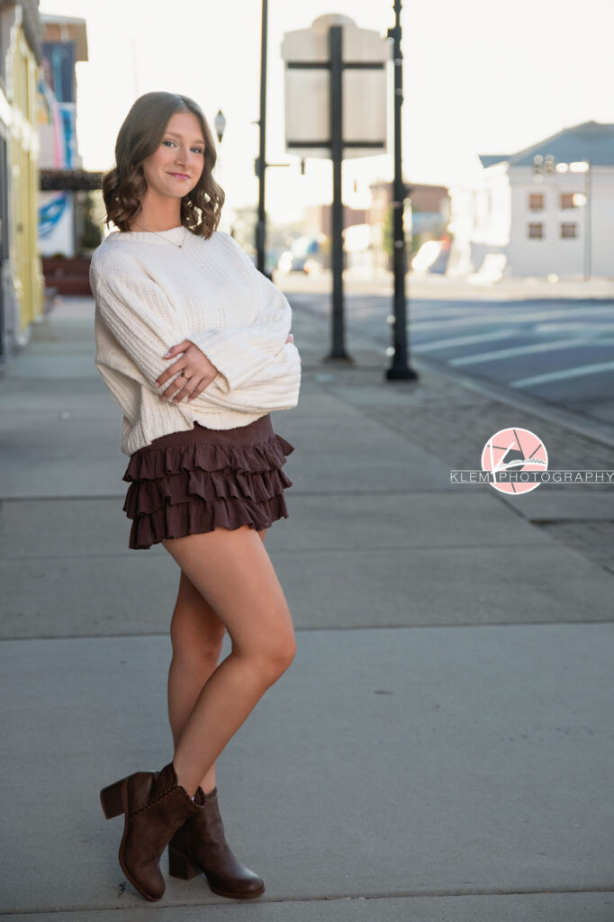Henderson KY Senior Pictures, Klem Photography. Full body shot of senior girl with short brown hair in curls and blue eyes, wearing white sweater, brown ruffled mini-skirt, and brown boots smiles at the camera. She stands with her arms crossed over her chest and the leg closest to the camera bent. There is a townscape in the background.