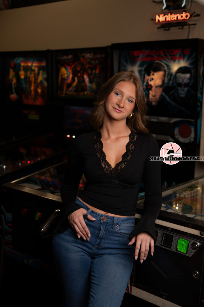 Henderson KY Senior Pictures, Klem Photography. Full body view of senior girl with short brown hair in curls and blue eyes, wearing a black long sleeve top and jeans smiles at the camera. She stands leaning against an arcade cabinet with one hand in her belt loop and the other on her arcade cabinet.