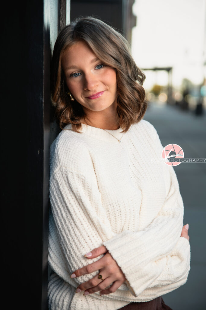 Henderson KY Senior Pictures, Klem Photography. Half body shot of senior girl with short brown hair in curls and blue eyes, wearing  a white sweater and rings smiles at the camera.  She stands with her arms crossed over her chest with a sidewalk visible in the background.