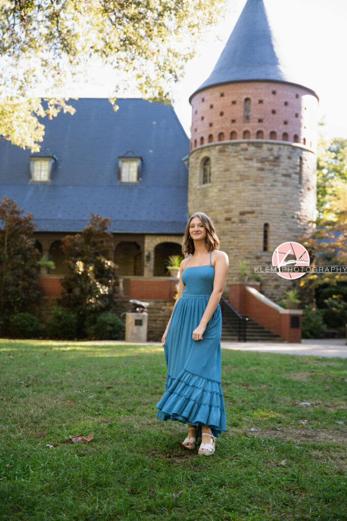 Henderson KY Senior Pictures, Klem Photography. Full body view of senior girl with short brown hair in curls and blue eyes, wearing long blue dress and white sandal heels looks at the camera. She stands with her arms by her sides with a castle and greenery in the background.