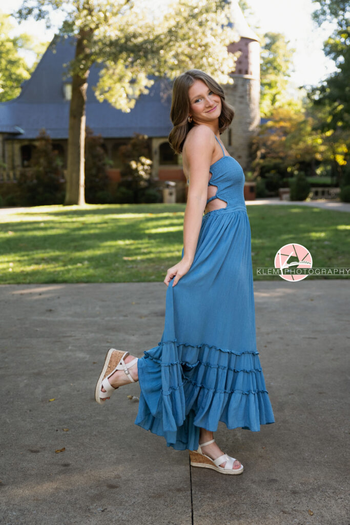 Henderson KY Senior Pictures, Klem Photography. Full body view of senior girl with short brown hair in curls and blue eyes, wearing a long blue dress and white sandal heels smiles at the camera. She stands with one leg up and one hand lifting up the bottom of her dress with a castle and greenery in the background.