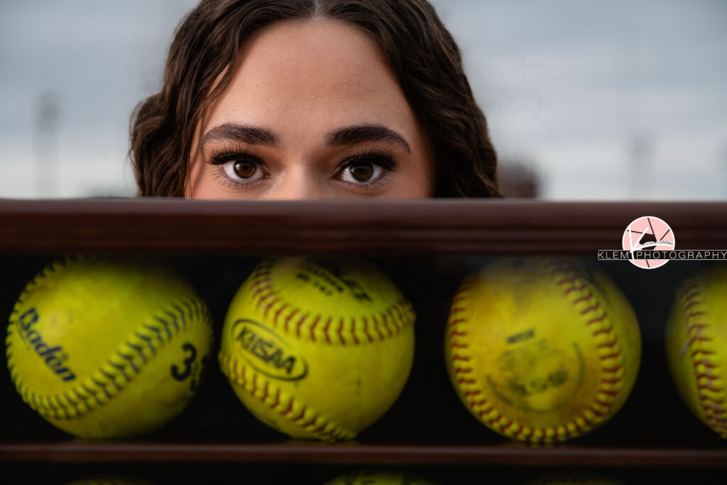 Henderson KY Senior Picture Ideas, Klem Photography, Mackenzie, Henderson County High School. Headshot of senior girl with long brown curly hair and brown eyes looks at the camera. Her eyes are clearly visible over a container of softballs.