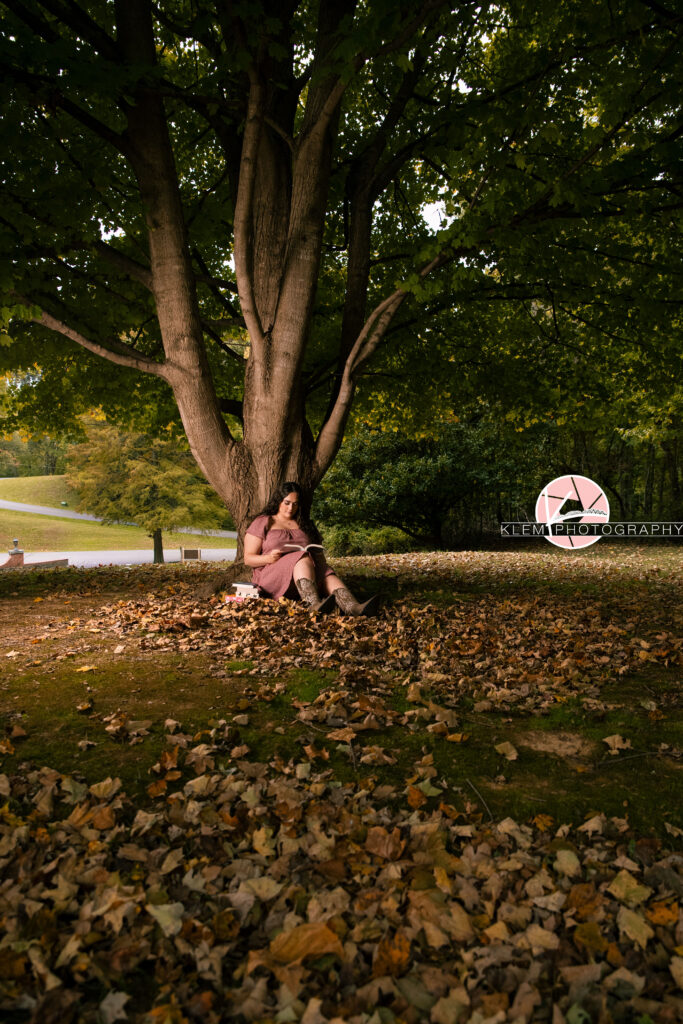 Henderson KY Senior Picture Ideas, Klem Photography, Mackenzie, Henderson County High School. Full body shot of senior girl with long brown curly hair wearing a red gingham dress and brown cowboy boots is reading a book. She sits under a tree with fallen leaves around her reading her book with more boots stacked to her side.